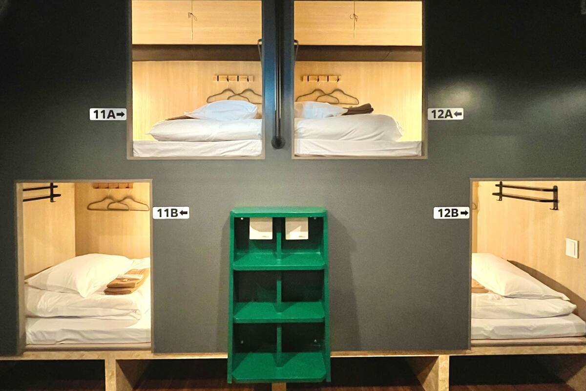 A room with four beds arranged in a square layout, each with a wooden headboard and white bedding. There are two small green storage units in the center.