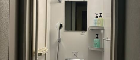 A small, enclosed shower area with a handheld showerhead, a wall-mounted mirror, and a shelf with toiletries.