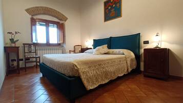 A bedroom with a bed, a window, a radiator, a chair, a table, and a lamp.