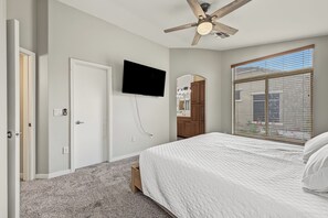 A bedroom with a bed, a ceiling fan, a TV mounted on the wall, a wardrobe, and a window with blinds.