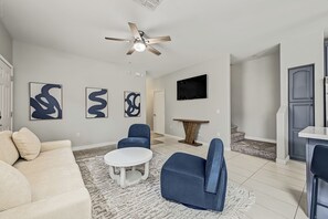 Een moderne woonkamer met een plafondventilator, abstracte wandkunst, een flatscreen televisie en een eethoek met een tafel en stoelen.