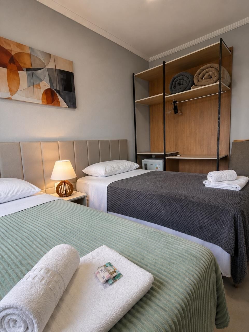 Hotellihuone, jossa on kaksi sänkyä, puinen hyllykkö ja taulu seinällä.
