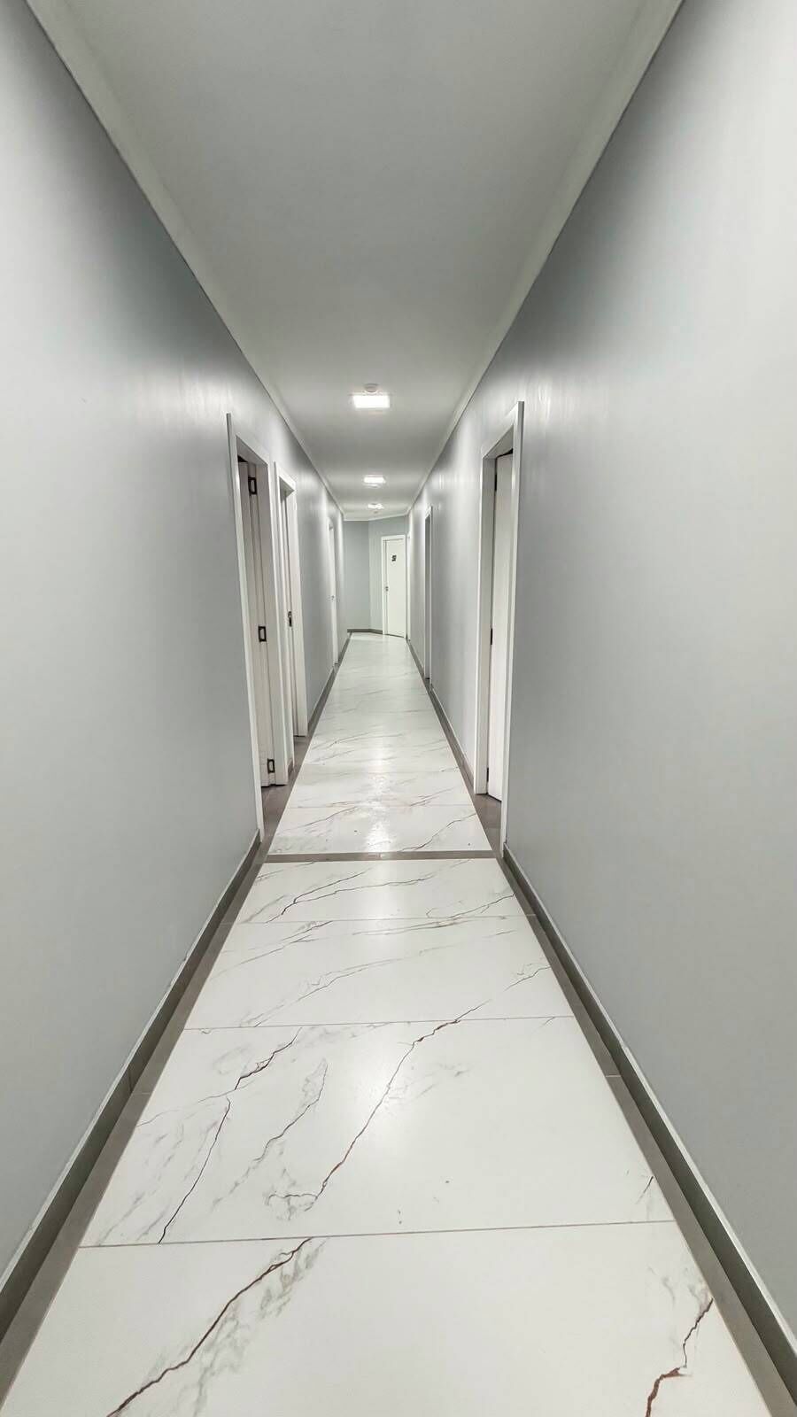 Un long couloir étroit aux murs blancs et au sol en marbre, qui mène à une porte au fond.