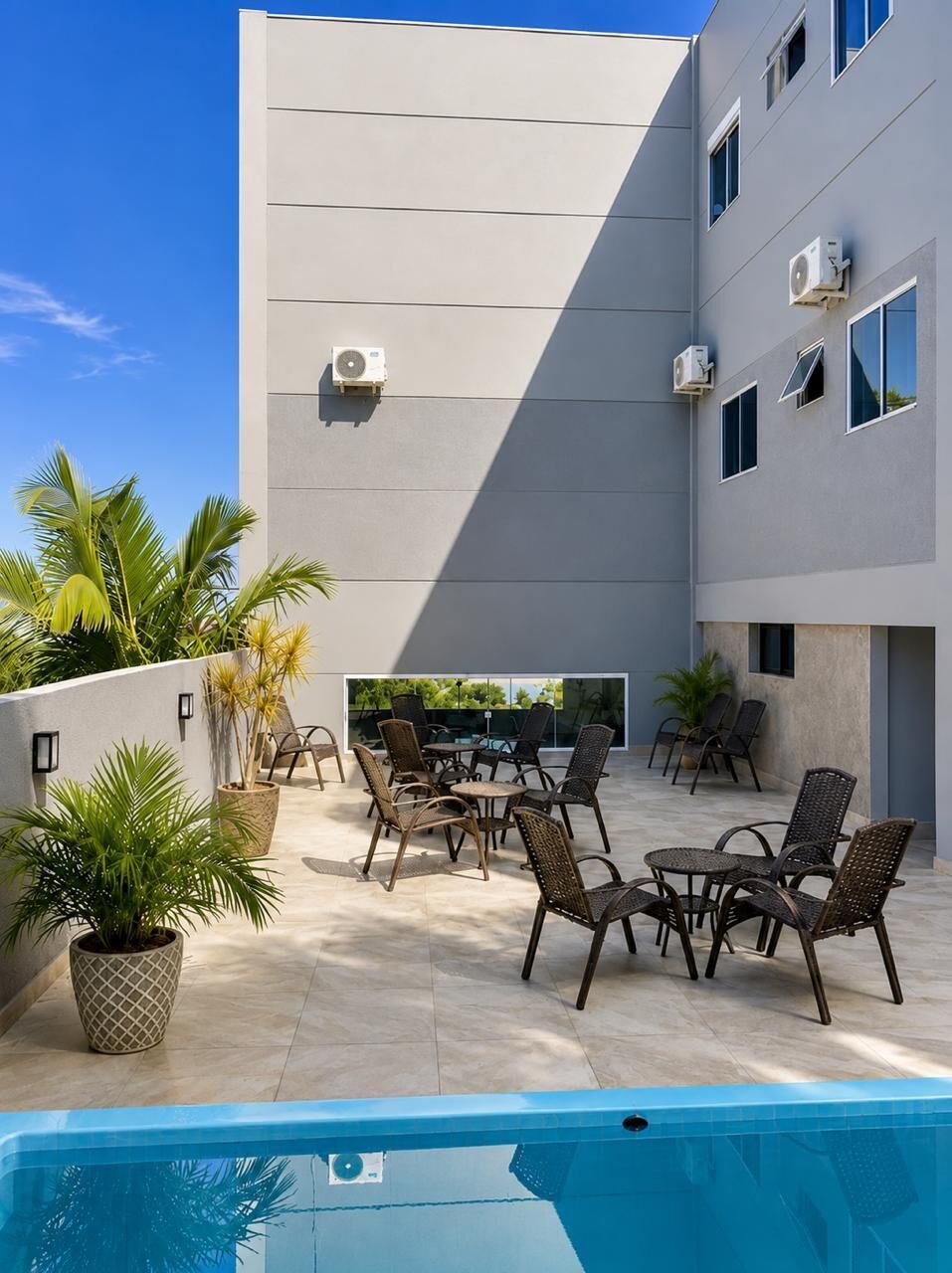 Zona de piscina al aire libre con sillones y mesas, rodeada por un edificio con unidades de aire acondicionado.
