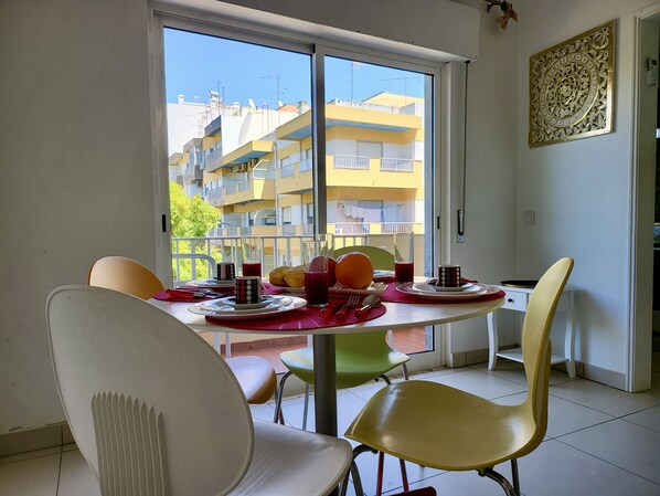 Un coin repas avec une table dressée pour quatre personnes, des chaises et une vue sur un immeuble d’habitation par une grande fenêtre.