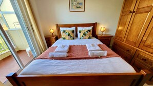 Una cama bien tendida con toallas, cabecera de madera, dos mesitas de noche con lámparas y vistas al balcón exterior.