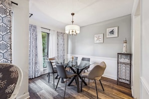 A dining area with a glass table, four chairs, a chandelier, patterned curtains, and a side table with a decorative item.