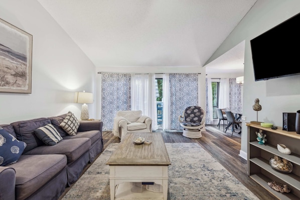 A living room with a sofa, coffee table, and TV. There is a dining area with chairs and a table in the background.