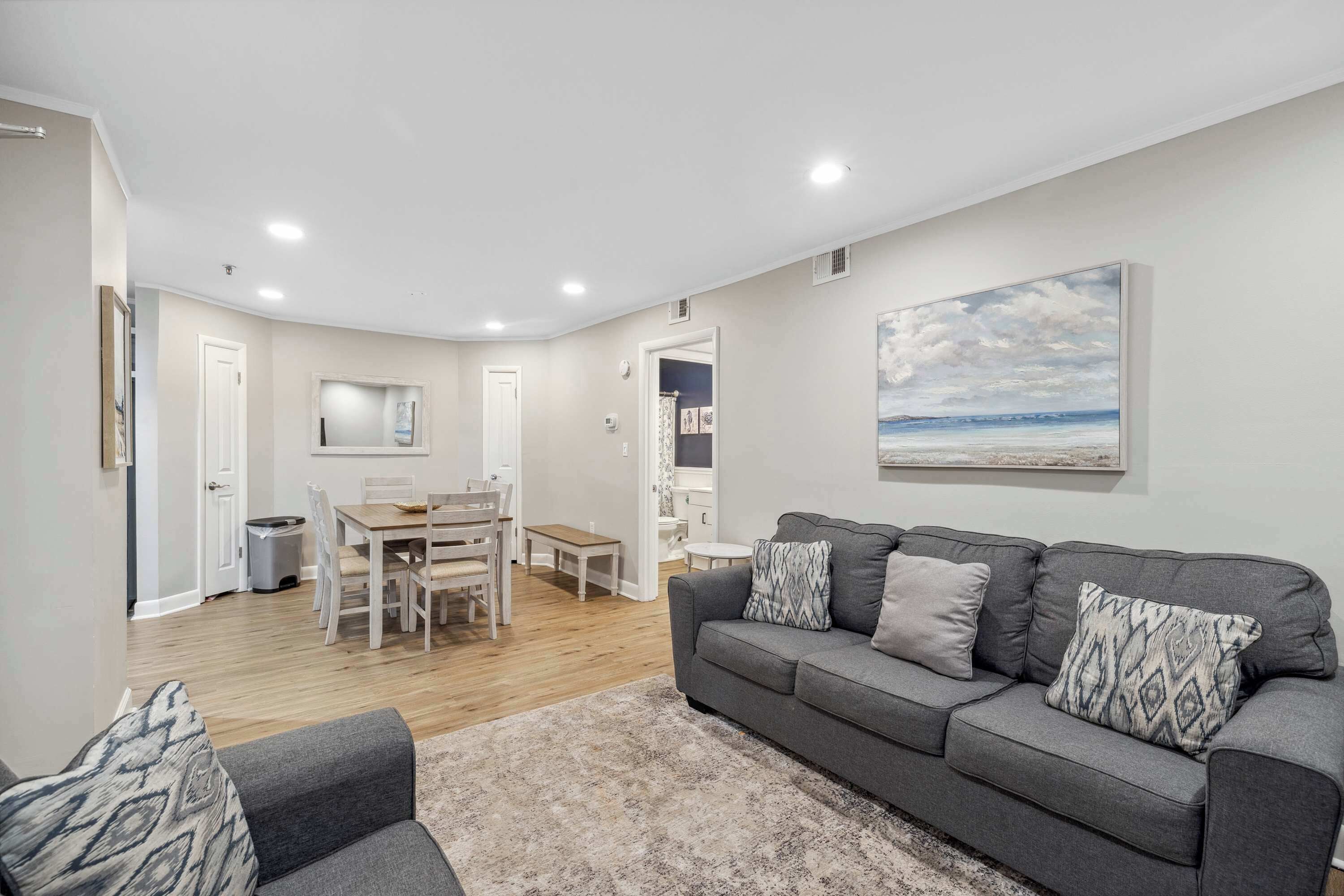 A modern living room with a grey sofa, a dining area with a wooden table and chairs, and a painting of a beach scene on the wall.