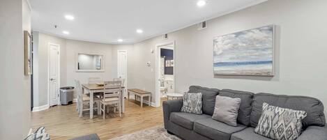 A modern living room with a grey sofa, a dining area with a wooden table and chairs, and a painting of a beach scene on the wall.