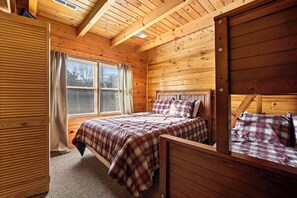 A wooden cabin bedroom with a bed, a window with curtains, and a bunk bed.
