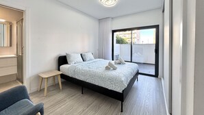 Une chambre moderne avec un grand lit, une petite table et une chaise. Une porte-fenêtre coulissante donne accès à un balcon.