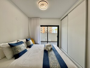A hotel room with a bed, a view of a building through a glass door, and a ceiling light. - Faro Prestige by Homing (Faro)