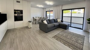 Un salon moderne avec un canapé d’angle gris, une table à manger avec des chaises, un coin cuisine équipé d’un four et une grande fenêtre donnant sur l’extérieur.