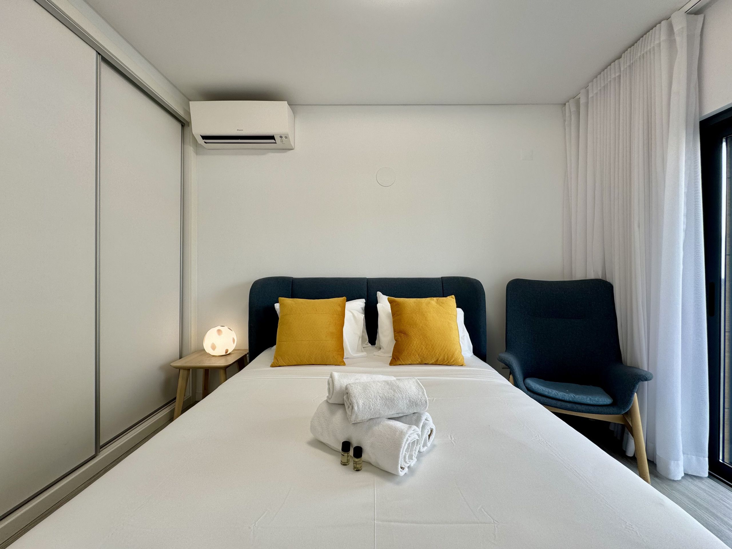 Una habitación de hotel con una cama, dos almohadas, una silla, una mesita de noche con lámpara y un equipo de aire acondicionado.