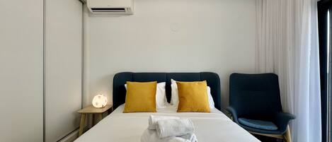 Een hotelkamer met een bed, twee kussens, een stoel, een nachtkastje met een lamp en een airconditioning.
