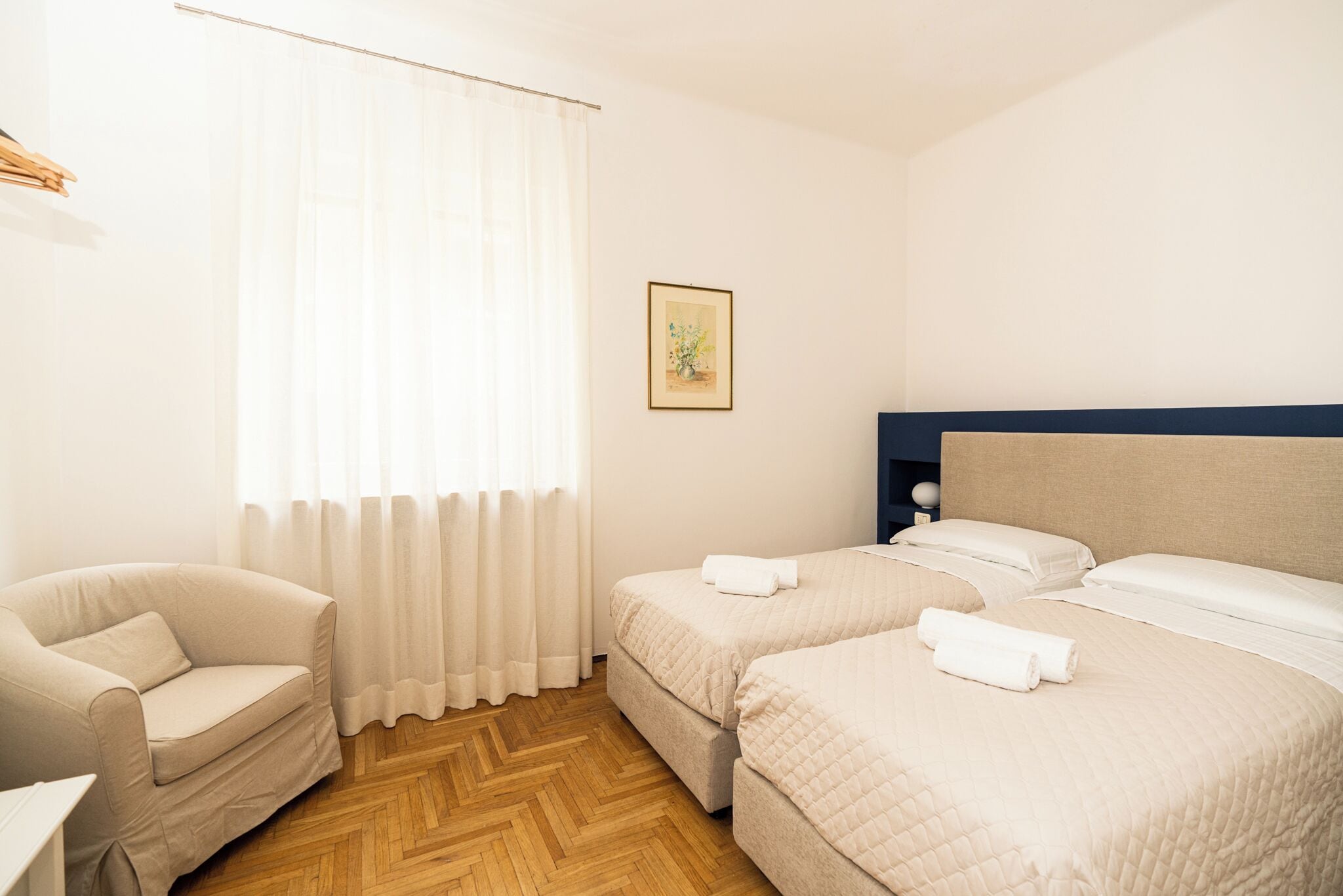 A hotel room with a wooden herringbone floor, two single beds with white linens, a beige armchair, and a large window with sheer curtains.