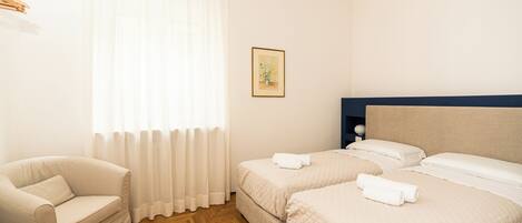 A hotel room with a wooden herringbone floor, two single beds with white linens, a beige armchair, and a large window with sheer curtains.