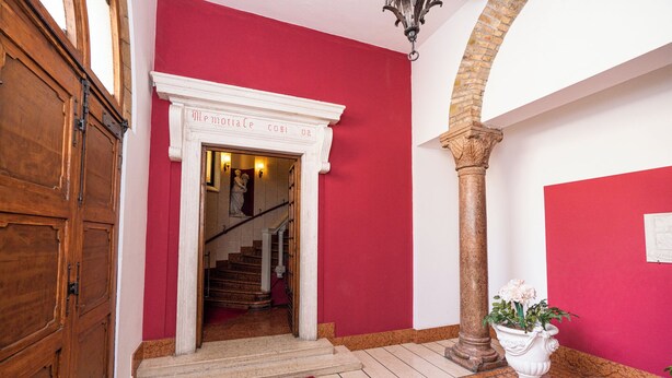 A red-walled hallway with a white door, a staircase, and a column. - Residenza Serego - affaccio scavi Romani (Verona)