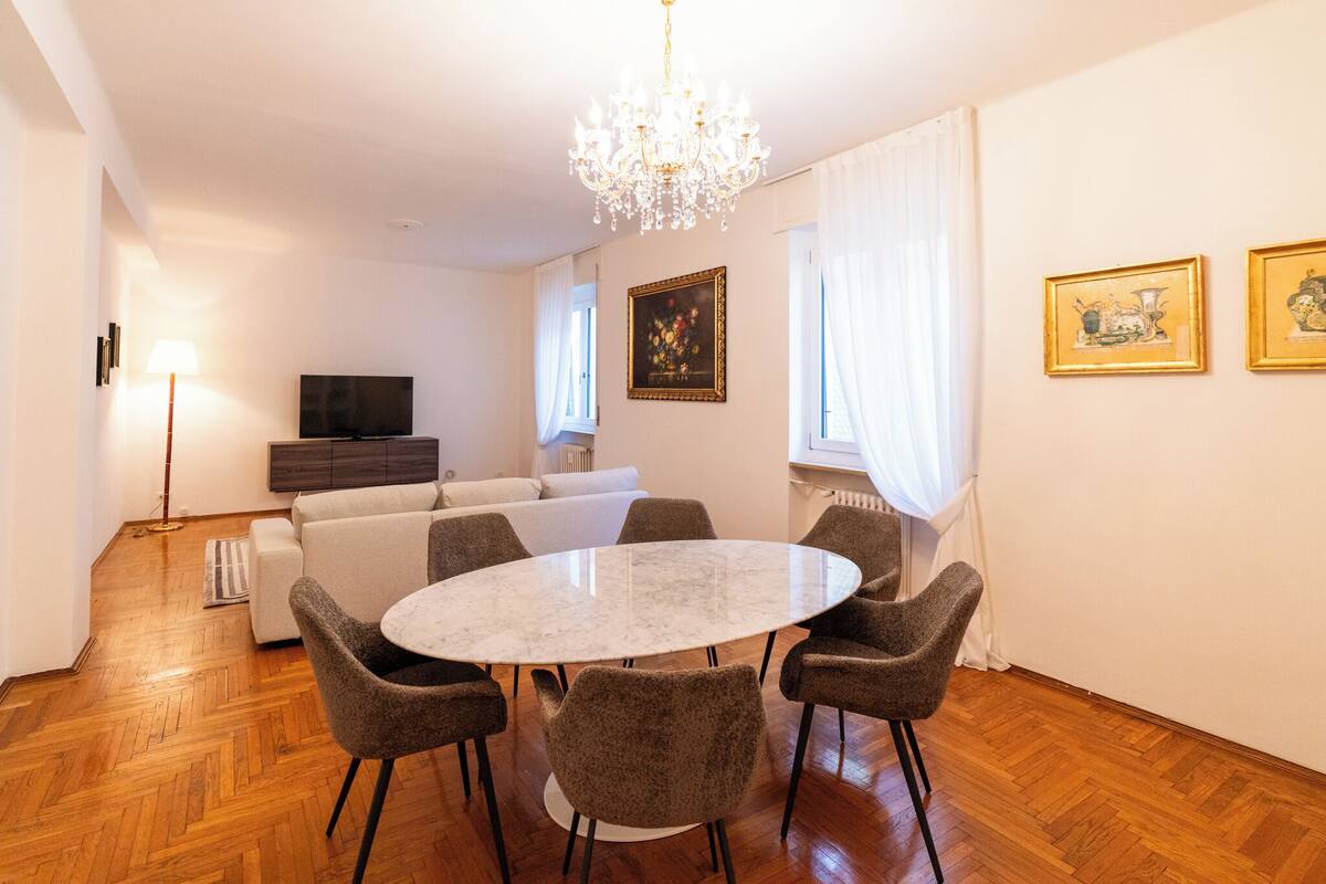 Uma área de refeições com uma mesa redonda e seis cadeiras, um sofá, uma TV e um lustre.