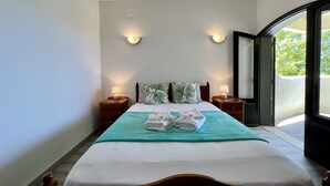 Une chambre d’hôtel comprenant un lit, deux tables de chevet avec des lampes, une fenêtre donnant sur un espace verdoyant et une porte menant à un espace extérieur.