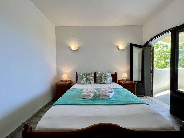A hotel room with a bed, two bedside tables with lamps, a window with a view of greenery, and a door leading to an outdoor area.