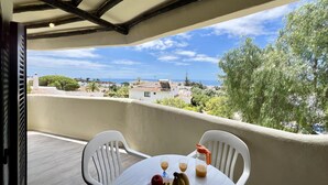 Un balcon avec une table dressée pour deux personnes, une bouteille de jus de fruits et une assiette de fruits. Le balcon offre une vue sur une ville côtière, avec ses bâtiments et ses arbres.