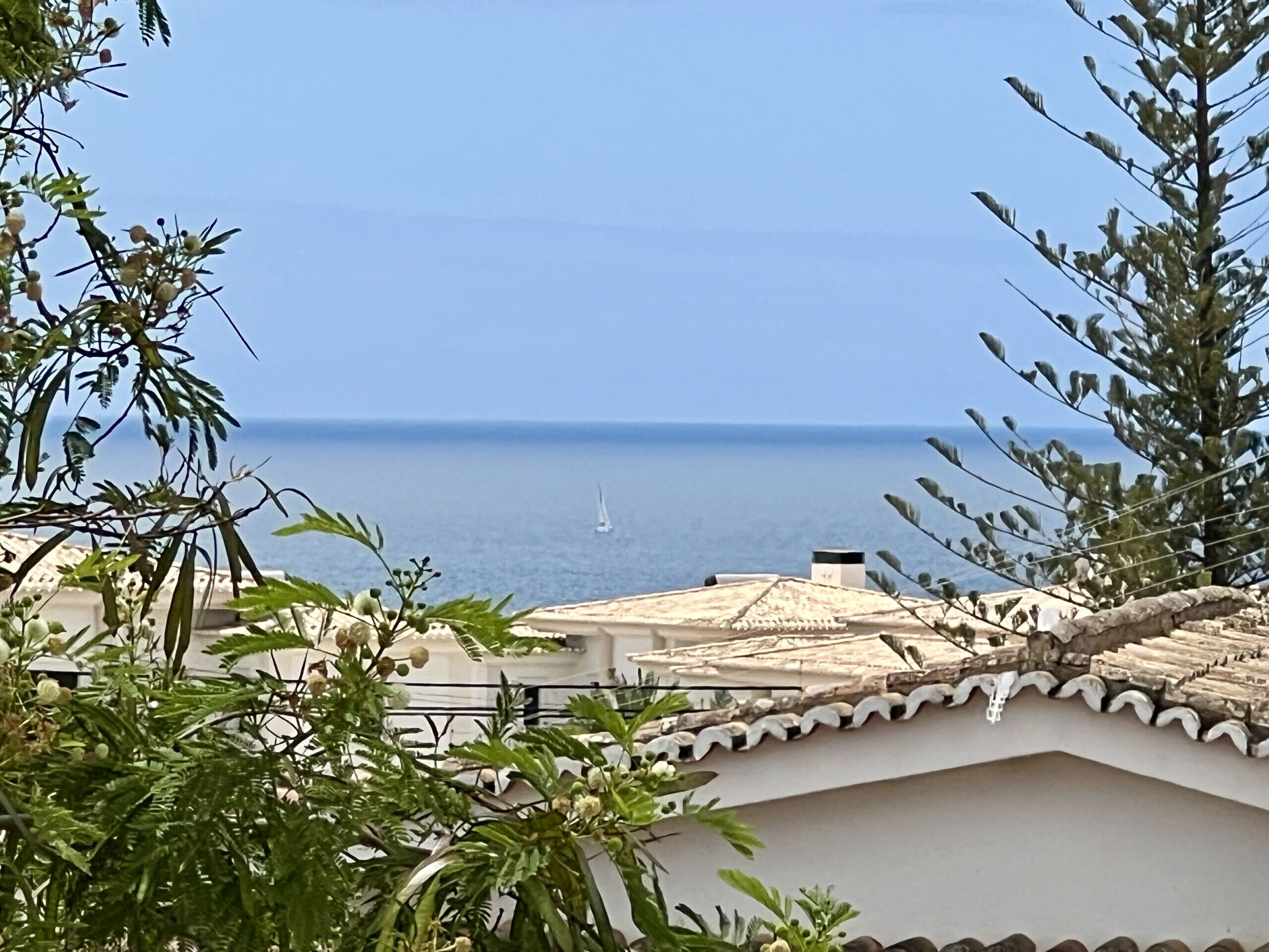 Une vue côtière avec un voilier sur la mer, entouré de verdure et de bâtiments à l’architecture traditionnelle.