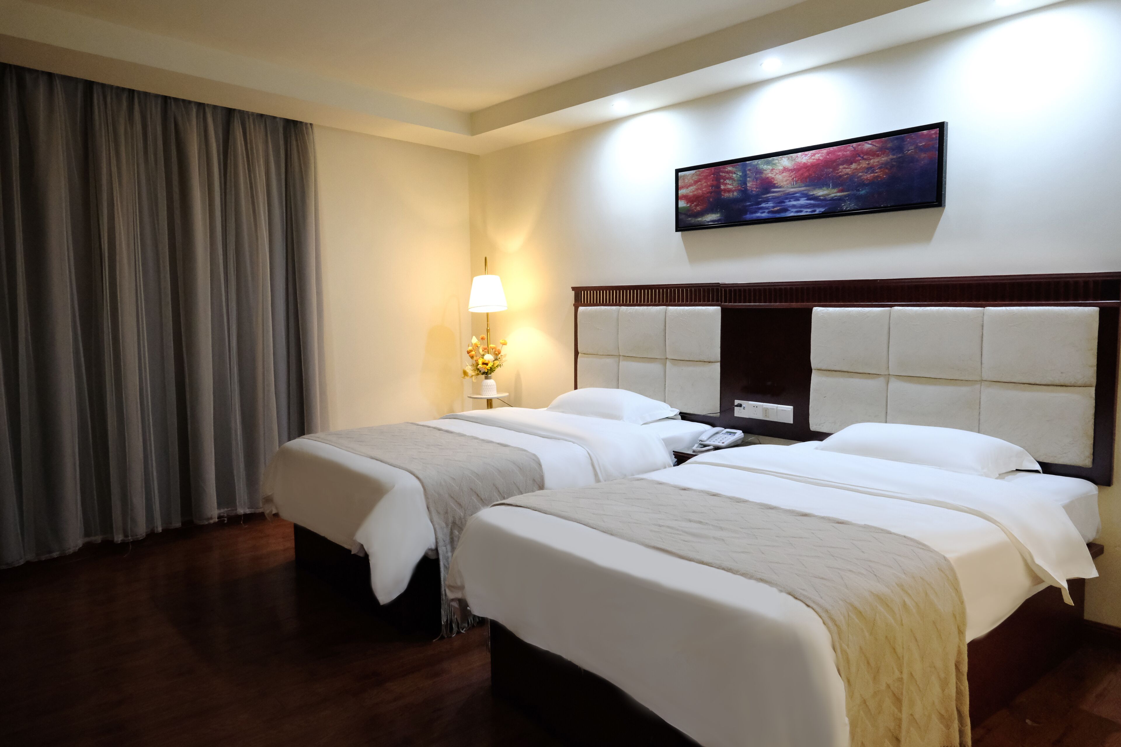 A hotel room with two beds, a nightstand, a lamp, a painting, and a window with curtains.