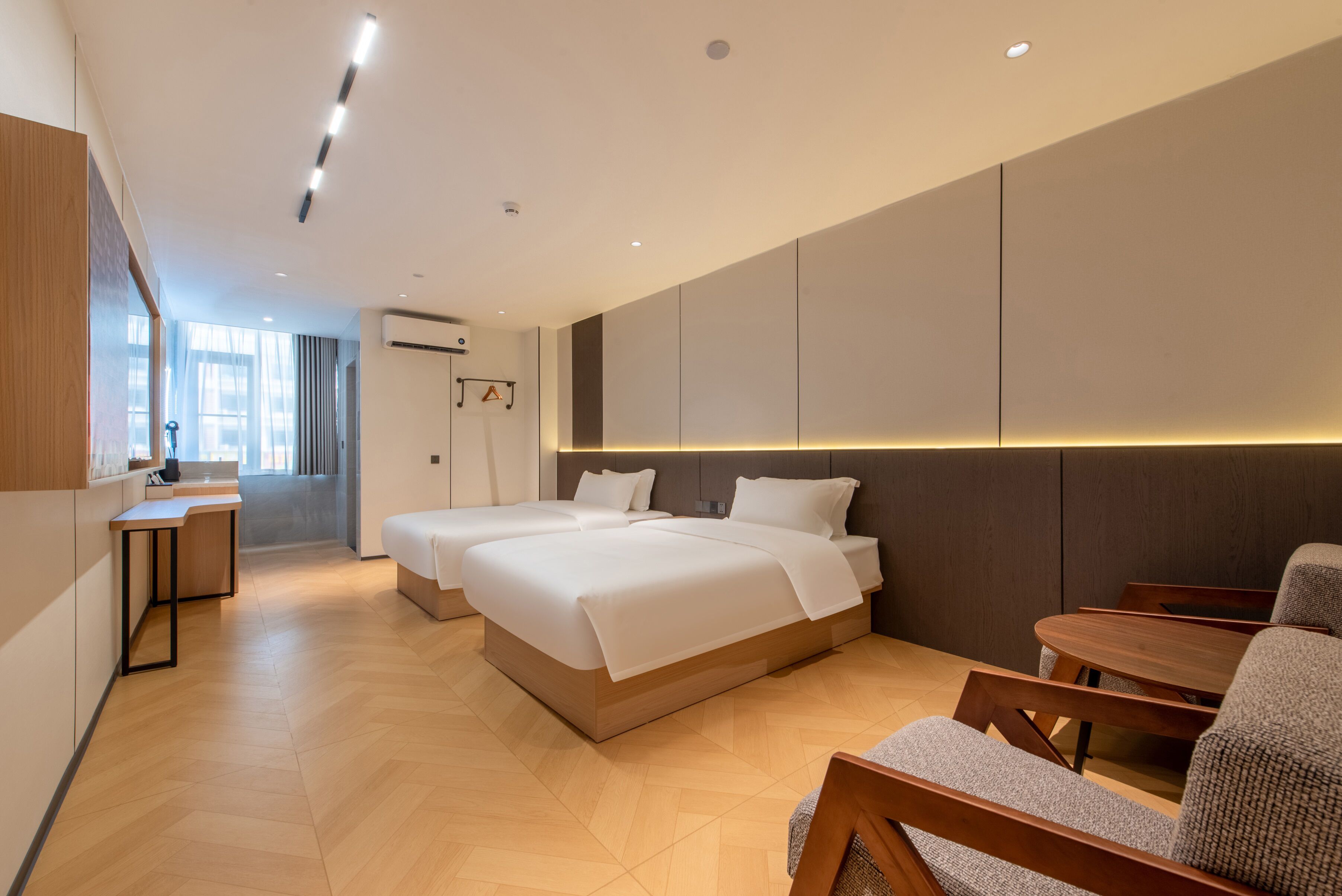 Une chambre d’hôtel moderne dotée de deux lits, d’un coin salon avec une table et des chaises, et d’une grande fenêtre laissant entrer la lumière naturelle.
