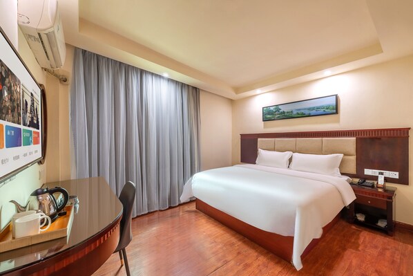 A hotel room with a large bed, a desk with a kettle, a chair, a wooden floor, a large window with curtains, a painting on the wall, and a nightstand with a phone.