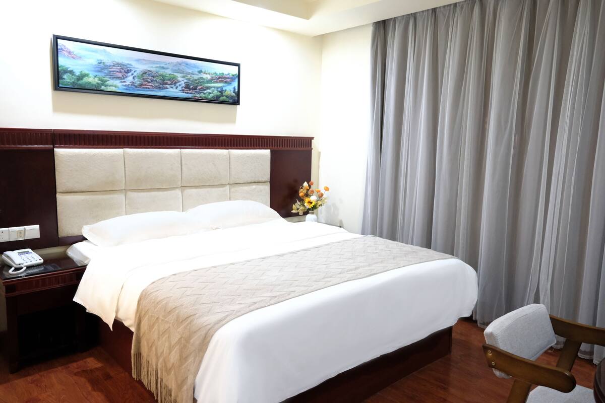 Een hotelkamer met een groot bed, een houten nachtkastje met een telefoon, een schilderij aan de muur en een raam met gordijnen.