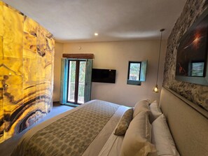 A bedroom with a bed, a TV mounted on the wall, a window with a view of greenery, and a stone wall. - Castello di Carlo - Suite Carlo IV (Val'Quirico)