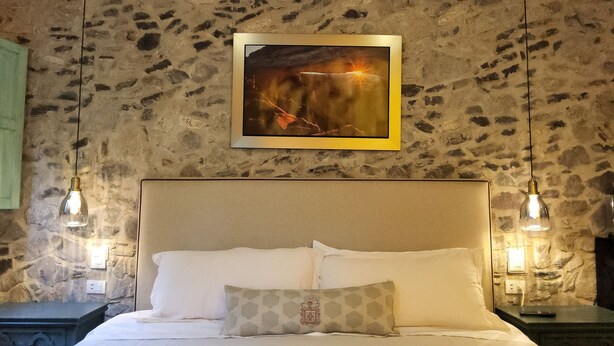 Una cama con sábanas blancas y un cojín estampado, una pared de piedra y dos lámparas colgantes.
