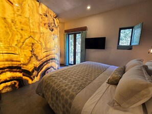A bedroom with a large, textured wall, a bed with patterned bedding, a television mounted on the wall, and a window with a view of greenery. - Castello di Carlo - Suite Carlo IV (Val'Quirico)