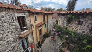 Una estrecha calle empedrada entre edificios de piedra con tejados de tejas rojas, una bandera y enredaderas con flores.