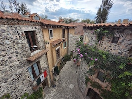 Una estrecha calle empedrada entre edificios de piedra con tejados de tejas rojas, una bandera y enredaderas con flores.