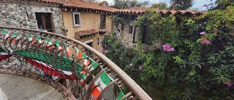 Un edificio de piedra con techo de tejas, baranda de hierro forjado con banderas y un jardín con plantas en flor.