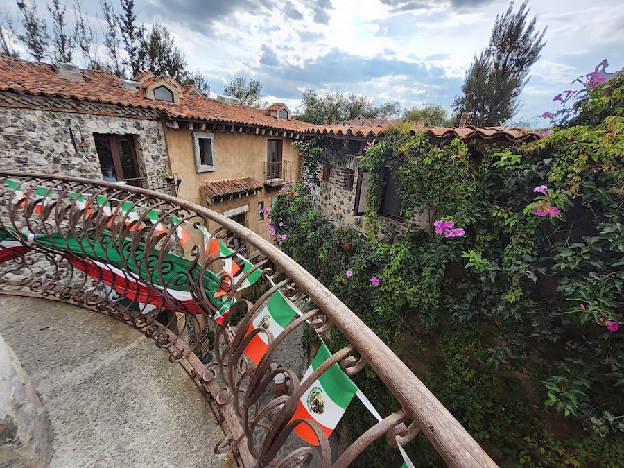 A stone building with a tiled roof, a wrought iron railing with flags, and a garden with flowering plants.