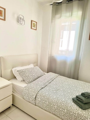 Quarto com cama, mesa de cabeceira, luminária na parede e janela com cortina.