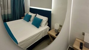 A hotel room with a large mirror, a bed with blue and white bedding, a bedside table with a lamp and remote control, and a nightstand with a lamp. - Luxurious Apartment with a sea view balcony in the N.Rhodes Yachting Marina Area (Rhodes)