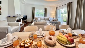 A well-lit living room with a sofa, a television, and a dining table set with breakfast items. - Vilamoura White House Villa by Homing (Loulé)