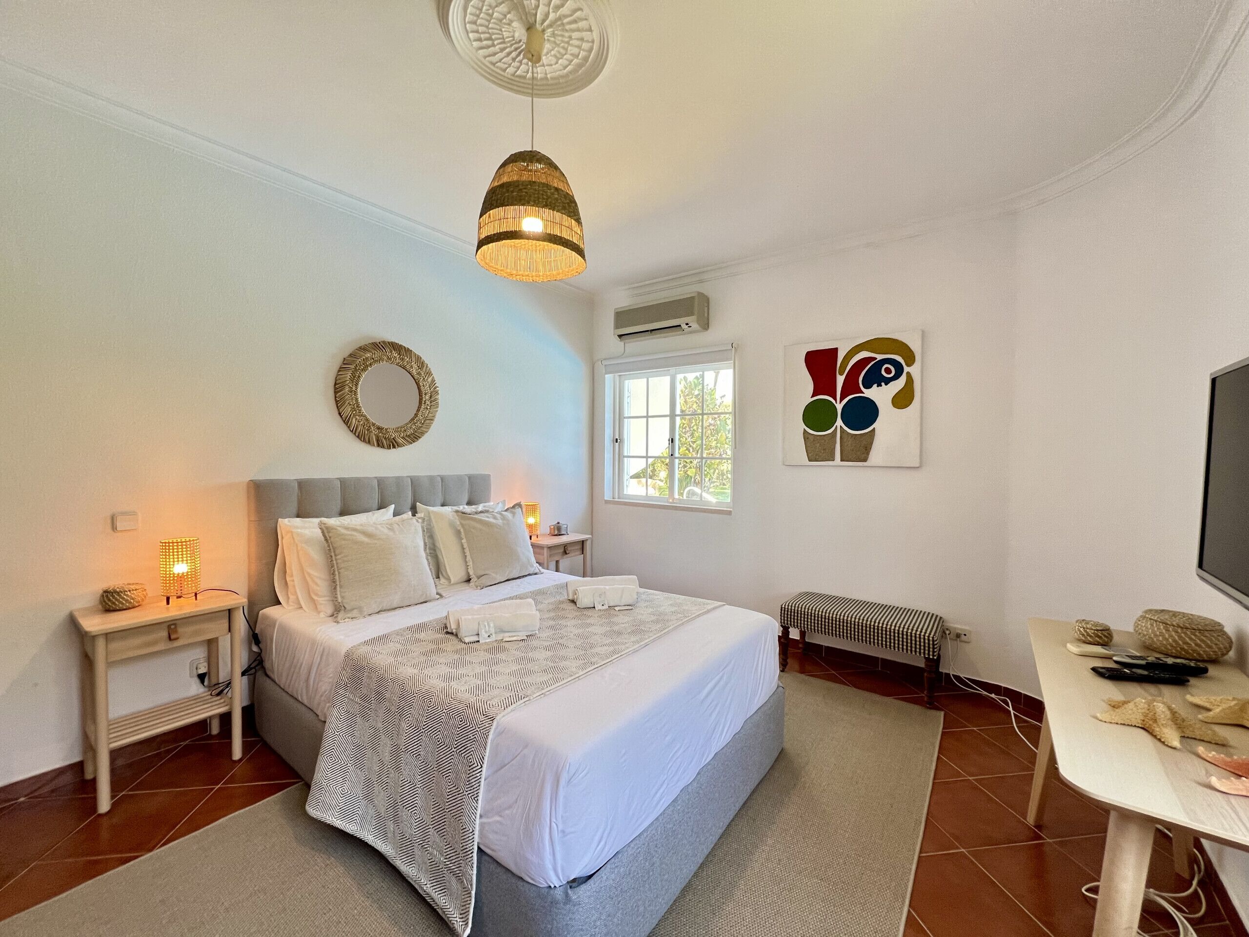 A bedroom with a bed, bedside lamps, a round mirror, a window, and a wall art piece.