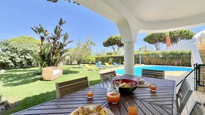 A patio with a table set for breakfast, including croissants and juice, overlooking a swimming pool and a well-maintained garden. - Vilamoura White House Villa by Homing (Loulé)