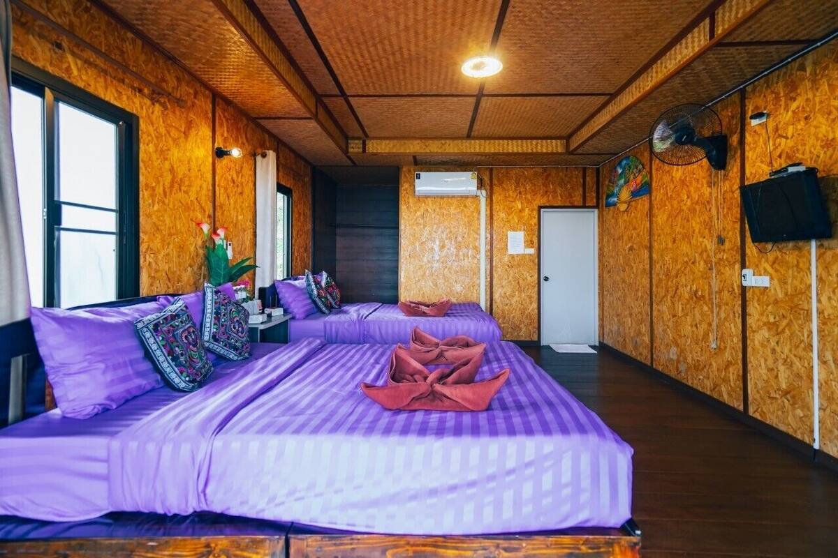 A room with wooden walls and a ceiling, a bed with purple bedding, a small table with a vase of flowers, and a door in the background.