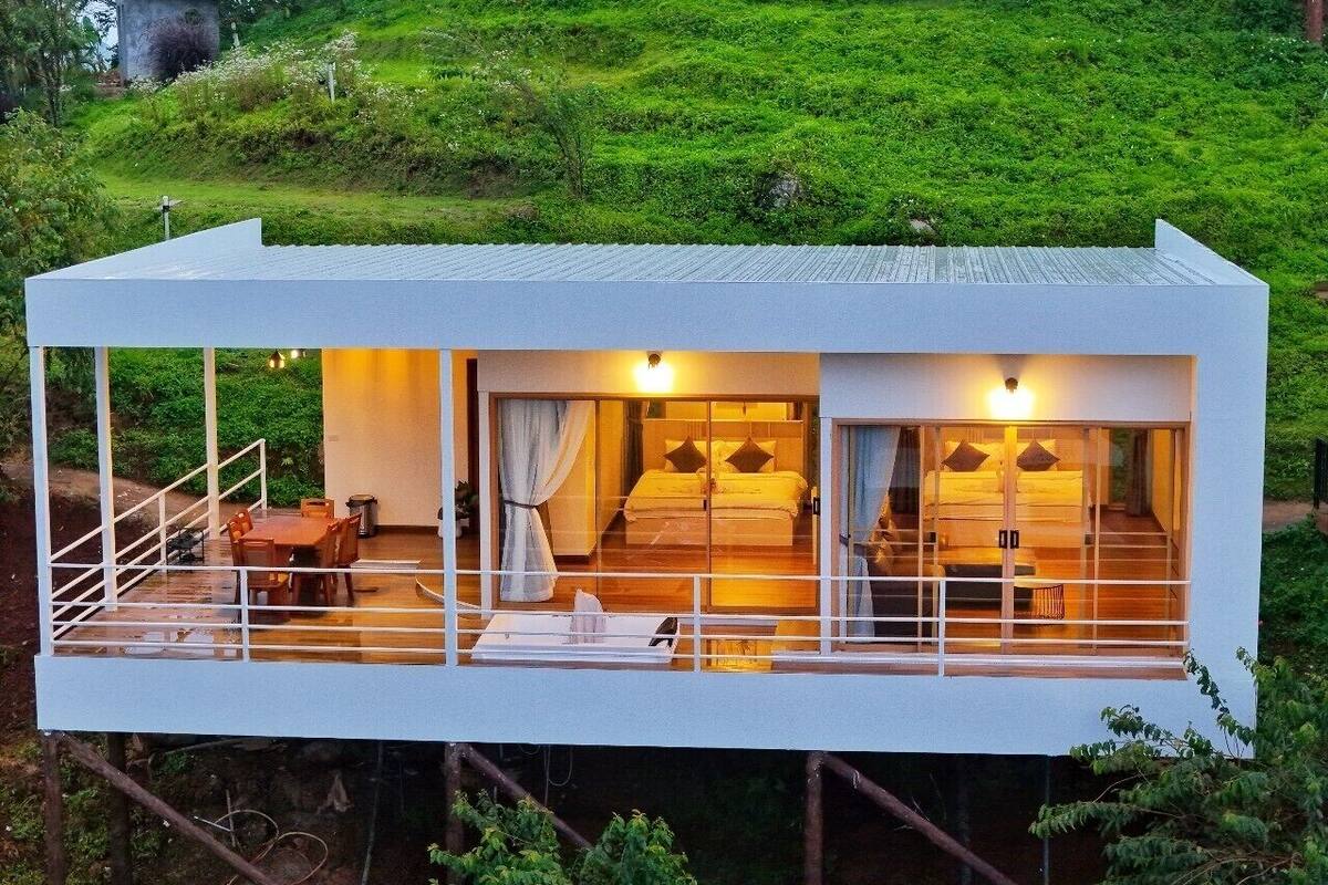 A modern, elevated guesthouse with a balcony, a dining area, and a bedroom with a large window.