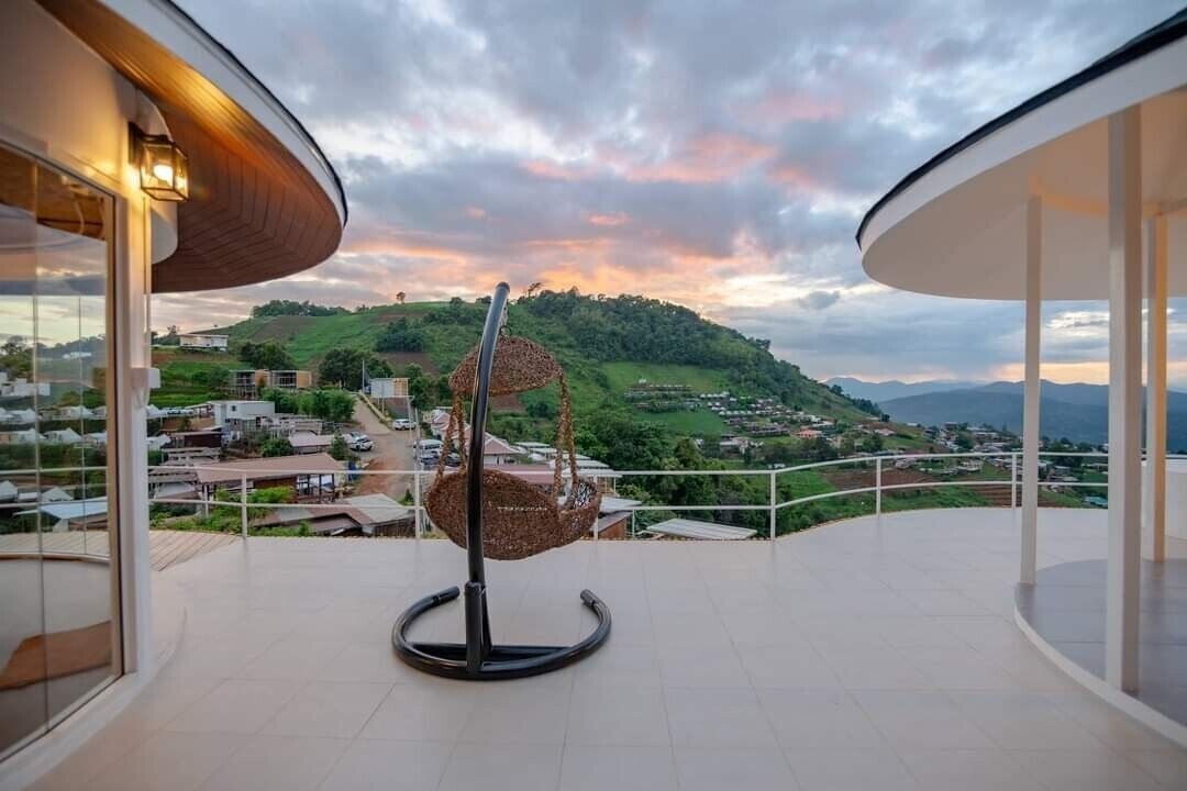 Un balcón con una silla colgante, vista a una ladera verde y un atardecer.
