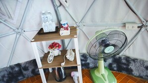 A room with a wooden shelf unit holding toiletries, a green fan, and a small table with a vase of flowers. - Mon Sai Lom Monjam (Mae Rim)