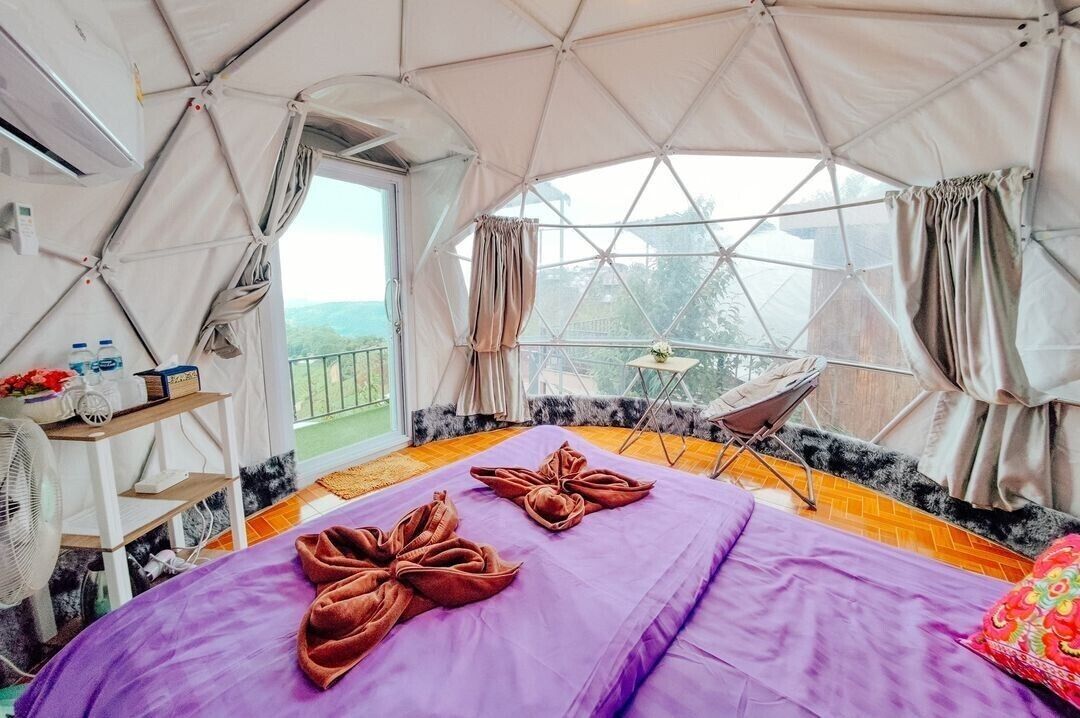 Habitación tipo tienda de campaña con una cama, un ventilador, una mesita con botellas y vistas al exterior.
