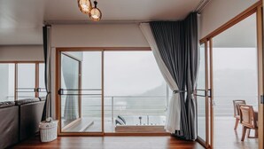 A modern room with a large sliding glass door leading to a balcony, a sofa, a small wicker basket, and a dining area with chairs. - Mon Sai Lom Monjam (Mae Rim)